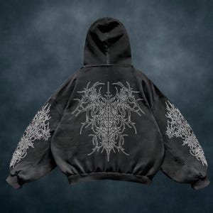 May include: Black hooded sweatshirt featuring a large white ornate design on the back and sleeves. The design is symmetrical and detailed, with a distressed, vintage appearance. The sweatshirt has a relaxed fit and a hood.
