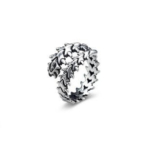 Può includere: Un anello color argento con un design dettagliato e strutturato. L'anello presenta un motivo ripetuto di forme stilizzate a forma di foglia, creando un accessorio unico e accattivante. L'anello è mostrato su sfondo bianco.
