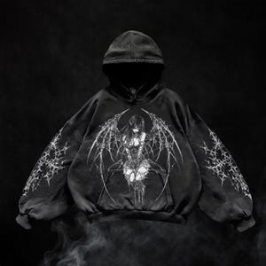May include: Black hooded sweatshirt featuring a detailed graphic of a woman with large, stylized wings. The design is in shades of gray and white, with intricate line work. The sleeves have a spiderweb-like pattern.