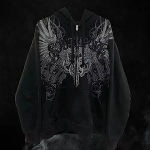 Peut inclure: Sweat à capuche noir zippé avec un motif graphique gris détaillé sur le devant. Le motif présente un design ailé avec des lettres ornées. Le sweat à capuche a une capuche et une poche avant. Le style général est audacieux et gothique.