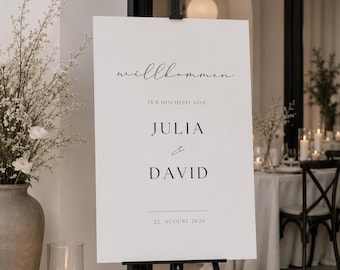 May include: A white rectangular wedding welcome sign displayed on a black metal tripod easel. The sign features black text that reads willkommen ZUR HOCHZEIT VON JULIA & DAVID 22. AUGUST 2026 in a mix of script and serif fonts with horizontal lines.