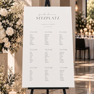 May include: A white wedding seating chart sign on a black easel, featuring the text "finde deinen SITZPLATZ", "MARINA & TOBIAS", "07. JULI 2027", and lists of names organized under nine tables labeled "TISCH 01" through "TISCH 09" in black font.