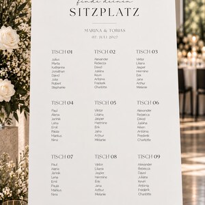May include: A white wedding seating chart sign on a black easel featuring the text finde deinen SITZPLATZ, MARINA & TOBIAS, 07. JULI 2027, and a list of names organized by table numbers from TISCH 01 to TISCH 09 in a clean black serif font.