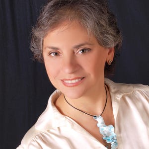 May include: A woman with grey hair is wearing a white blouse with a blue and white glass pendant necklace.