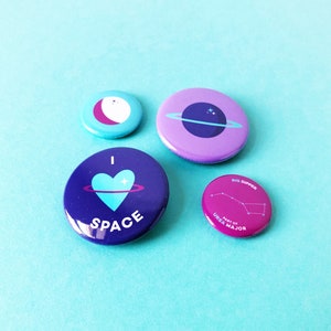 May include: Four colourful space-themed badges. One badge has a crescent moon design, another features a planet with rings, and a third says "I heart SPACE" with a heart and planet graphic. The last badge shows the Big Dipper constellation.