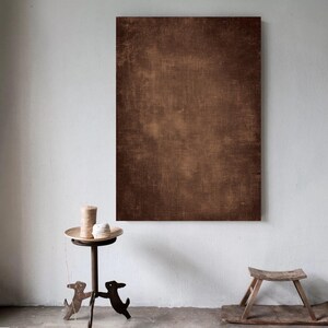 May include: A large, rectangular brown canvas with a textured surface hangs on a white wall. A small, decorative table with a round top and dog-shaped legs holds a candle and a ball of twine. A small wooden stool sits on the floor.