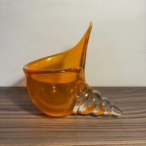May include: An orange glass seashell-shaped decorative bowl. The bowl has a curved body and a pointed spout. The base of the bowl is a clear, spiraled design. The bowl is sitting on a wooden surface.