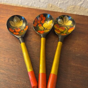 May include: Three wooden spoons with black bowls and yellow handles. The bowls are decorated with floral patterns in yellow, orange, and red. The ends of the handles are painted red.