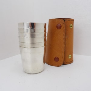 May include: A set of six silver metal cups and a brown leather carrying case with snap closures. The cups are stacked and have a shiny, reflective surface. The leather case is cylindrical.