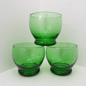 May include: Three green glass tumblers. The glasses have a rounded, bulbous shape and a thick, rounded base. The glasses are a vibrant green colour and appear to be vintage.