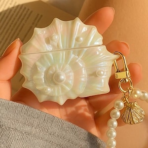 May include: A pearlescent white seashell-shaped earbud case, held in a hand. The case has a gold-coloured clasp and is attached to a pearl bracelet with a seashell charm. An open book and a person's legs are in the background.