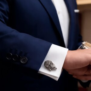 Peut inclure: Un bouton de manchette en métal argenté en forme de vaisseau spatial avec des détails mécaniques complexes, fixé sur la manchette blanche d'une chemise à manches longues portée sous un blazer bleu marine foncé avec des boutons assortis.
