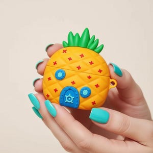 May include: A yellow pineapple-shaped case with a green top and red accents. The case has a blue door and windows. The case is designed to hold earbuds.
