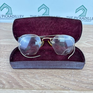 May include: Vintage eyeglasses with a gold-tone metal frame and clear lenses rest inside a maroon velvet-lined case. The glasses have a unique, angular shape. The case is open, revealing the glasses against the plush interior.