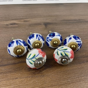 May include: Six ceramic cabinet knobs with floral designs. The knobs have a white base with blue, red, and green floral patterns. Each knob has a gold or silver-colored metal base and stem.