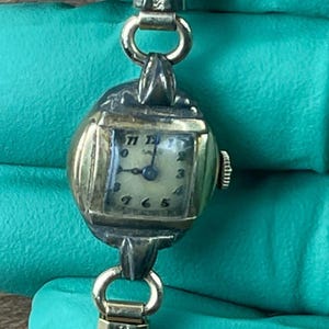 May include: Vintage gold-toned wristwatch with a rectangular face and a cream-colored dial. The watch features black numerals and hands, and is connected to a gold-toned chain. The watch is an oval shape and has a crown on the side.