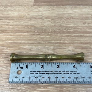 May include: A brass-colored, cylindrical object with flared ends and a central, decorative band. A ruler with inch markings is placed next to the object. The ruler's text provides instructions for reading measurements in centimeters and millimeters.