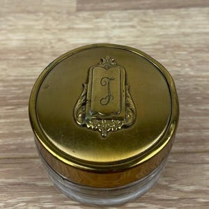 May include: A vintage glass jar with a brass-colored lid. The lid features an ornate design with a central rectangular cartouche bearing the letter "J" in a script font. The jar's clear glass body is visible below the lid.
