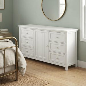 May include: A white wooden dresser featuring six drawers and a central cabinet door with vertical paneling. Each drawer and the cabinet door are equipped with a single round white knob. The dresser has a rectangular top and a flat base.