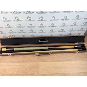 May include: A wooden pool cue set in a brown case. The case is open, revealing a cue stick with a green grip and a second stick. The case has a black interior and gold-colored latches. The word "IMPERIAL" is visible.