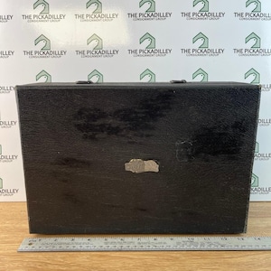 May include: A black, rectangular box with a textured surface. A small, silver-colored metal plate is affixed to the front. The box has a hinged lid and a metal clasp. A ruler is at the bottom.