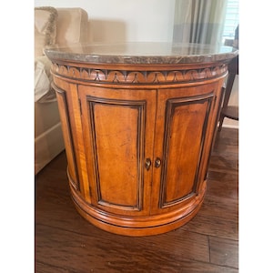 May include: A round, wooden cabinet with two doors and a marble top. The cabinet is a warm, medium brown color with darker brown trim. The top is a mottled brown and gray. The cabinet has a decorative carved design around the top.
