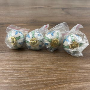 May include: Four decorative cabinet knobs. Each knob is white with blue and green floral designs and a gold-colored center. The knobs are individually wrapped in clear plastic and are displayed on a wooden surface.