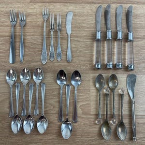 May include: An assortment of vintage silverware, including forks, knives, and spoons, arranged on a wooden surface. The collection features various designs, from simple to ornate, with clear acrylic handles on some knives.