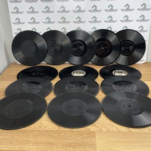 May include: A collection of twelve black circular vinyl records arranged in four rows. Each record features a central label with text, including the Edison brand name. The records are smooth, glossy, and uniform in shape, displayed on a flat surface.