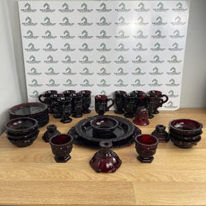 May include: A collection of deep red pressed glass tableware featuring a repeating geometric pattern. The set includes multiple mugs, small bowls, plates, candle holders, and a bell, all displaying a consistent dark ruby hue and textured finish.