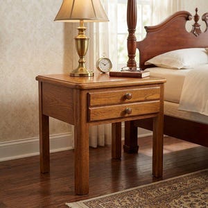 May include: A square wooden nightstand featuring two stacked drawers with small round knobs. The piece is constructed from medium-toned oak wood with a visible grain pattern, supported by four straight legs, and positioned next to a bed.