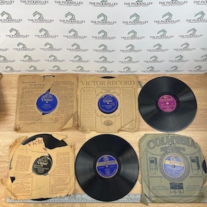May include: A collection of vintage vinyl records and their sleeves. The records are black with labels in various colors, including blue and purple. Some sleeves are tan with printed text and logos, including "Victor" and "Columbia".