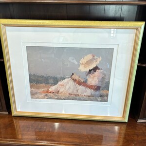 May include: A framed artwork featuring an impressionistic painting of a person wearing a hat and white dress. The painting has a soft color palette of beige, white, and brown. The frame is gold with a white mat.