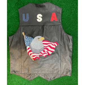May include: A black leather vest featuring the letters U, S, and A in blue, white, and red across the upper back. Below the text is an embroidered patch depicting a bald eagle with a yellow beak positioned in front of a waving American flag.