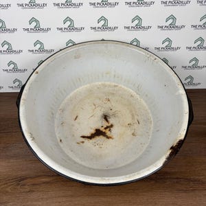 May include: A vintage white enamelware pot with a black rim and handles. The interior shows signs of wear and discoloration, with a dark brown stain at the bottom. The pot is round and has a wide opening, suitable for cooking or storage.