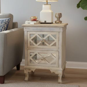 May include: A light-colored wooden nightstand featuring two drawers with mirrored panels and a decorative diamond-shaped lattice overlay. Each drawer is equipped with a clear, faceted crystal-style knob. The piece stands on four curved legs.