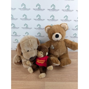 May include: Three plush toys: a tan dog, a brown teddy bear, and a Curious George monkey wearing a red shirt. The teddy bear has a silver medallion. The background features a repeating logo.