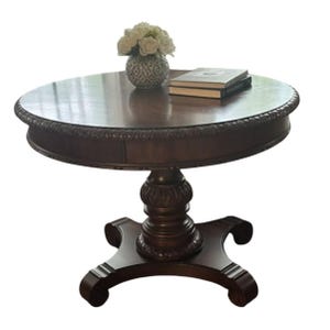 May include: A round dark brown wooden pedestal table featuring a carved edge detail along the tabletop, a central column base with decorative molding, and four curved feet. A patterned vase with white flowers and two books sit on top.