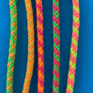 May include: Close-up of several colourful braided cords in neon green, orange, and pink. The cords are vertically aligned against a solid blue background. The cords have a textured appearance, with the colours interwoven to create a patterned effect.