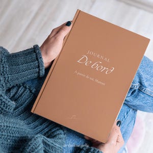May include: A brown journal with the words "JOURNAL De bord" and "A partir de toi, Maman" in white script. The journal is held by a person wearing a blue sweater and jeans. A small musical note is at the bottom.
