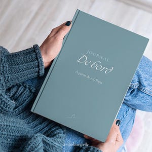 May include: A teal-colored journal with the words "JOURNAL De bord" and "À partir de toi, Papa" printed on the cover. The journal is held in a person's hands, with a blue sweater and jeans visible in the background. A small musical note is at the bottom.