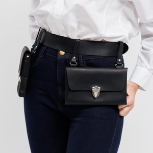 May include: Black leather belt bag with a rectangular main compartment and a smaller side pouch. The bag features a silver-colored clasp and is worn with a black belt and suspender straps. The person is wearing dark blue jeans and a white shirt.
