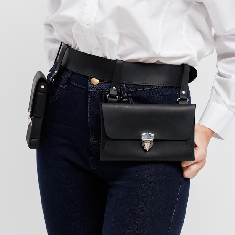 May include: Black leather belt bag with a rectangular main compartment and a smaller side pouch. The bag features a silver-colored clasp and is worn with a black belt and suspender straps. The person is wearing dark blue jeans and a white shirt.