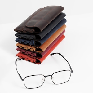Pode incluir: Uma pilha de seis estojos de &oacute;culos de couro em v&aacute;rias cores: castanho escuro, preto, azul marinho, bege, bord&ocirc; e vermelho. Um par de &oacute;culos com arma&ccedil;&atilde;o preta est&aacute; em frente aos estojos.