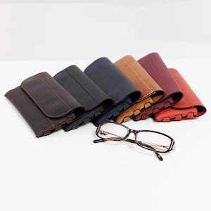 Pode incluir: Uma cole&ccedil;&atilde;o de estojos de &oacute;culos de couro em v&aacute;rias cores, incluindo castanho, preto, azul marinho, bege, bord&ocirc; e vermelho. Um par de &oacute;culos com arma&ccedil;&atilde;o tartaruga est&aacute; em frente aos estojos.