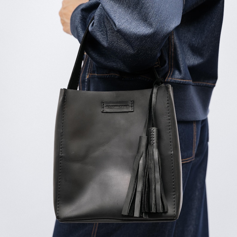 K&ouml;nnte beinhalten: Schwarze Ledertasche mit rechteckiger Form und langem Schulterriemen. Die Tasche hat einen dekorativen Quastenanh&auml;nger und Ziern&auml;hte. Der Markenname ist auf einem kleinen rechteckigen Aufn&auml;her sichtbar.