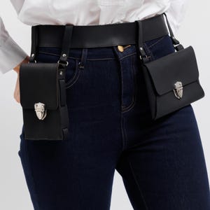 May include: Black leather belt with two small, matching black pouches. Each pouch has a silver-colored clasp. The belt is worn with dark blue jeans and a white shirt. The pouches are attached to the belt with straps.