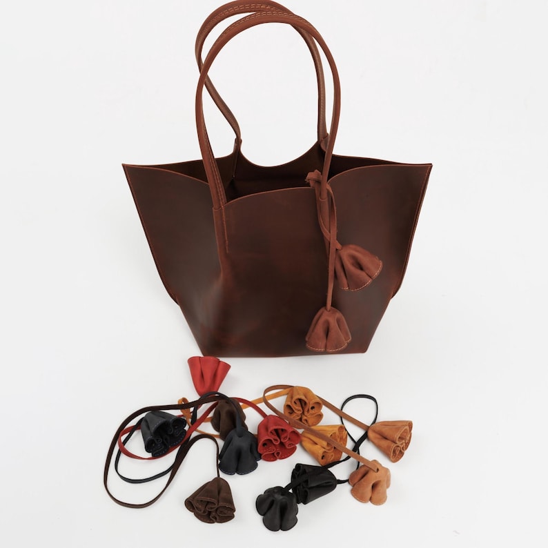 May include: A brown leather tote bag with matching handles and decorative tassels. Several colorful leather flower-shaped charms are scattered below the bag. The bag has a simple, elegant design.