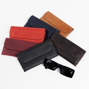 Pode incluir: Uma variedade de estojos de &oacute;culos de couro em v&aacute;rias cores, incluindo vermelho, preto, castanho e azul marinho. Os estojos retangulares t&ecirc;m fecho de aba e detalhes costurados. Um par de &oacute;culos de sol pretos est&aacute; ao lado.