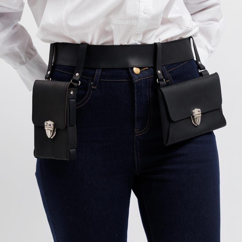 May include: Black leather belt with two attached pouches. Each pouch has a flap closure with a silver-colored clasp. The belt is worn with dark blue jeans and a white shirt. The pouches are rectangular and hang from the belt.